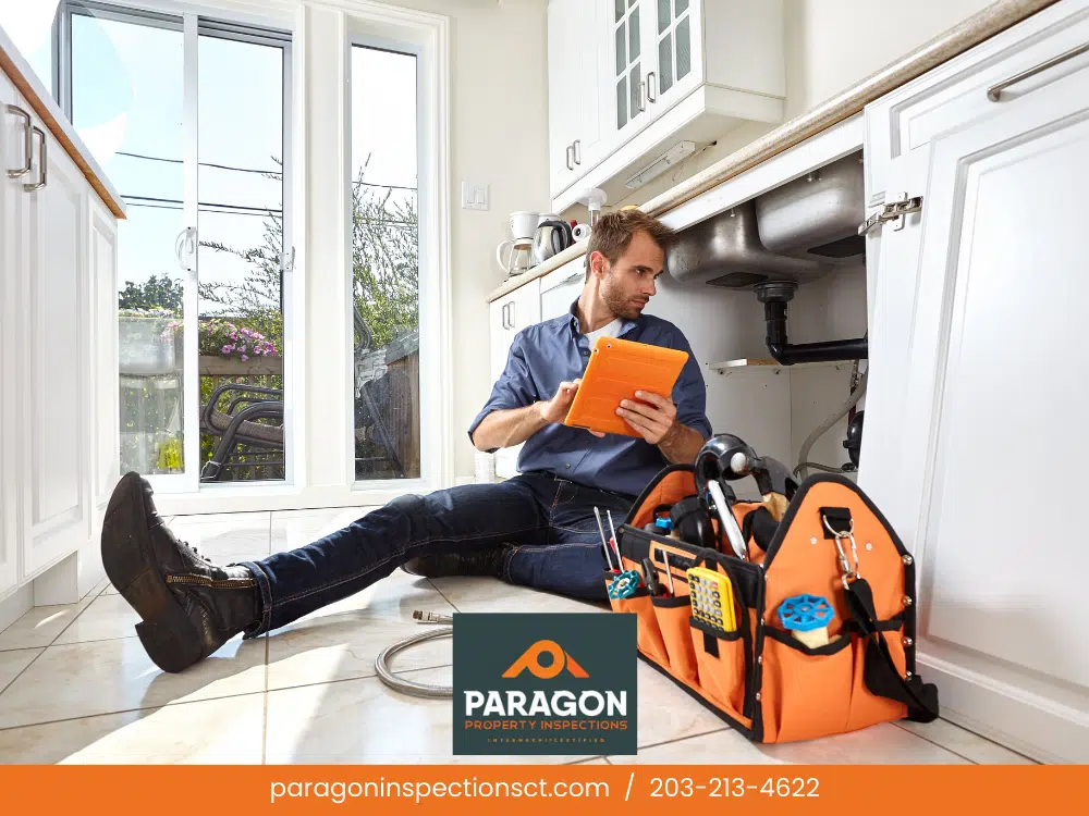 Mid-Year Plumbing Tip from Paragon Property Inspections: Save Water & Avoid Costly Repairs. A home inspector sits on the kitchen floor, inspecting under the sink with a tablet, surrounded by tools in a bag.